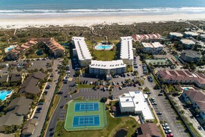 An aerial view of a coastal resort with multiple buildings, a central pool, tennis courts, and a beach in the background.