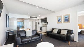A modern living room with two black leather sofas, a white coffee table, and a kitchen in the background. - TOP FLOOR H Residence 13204 - Level 32 2 bedrooms (Surfers Paradise)