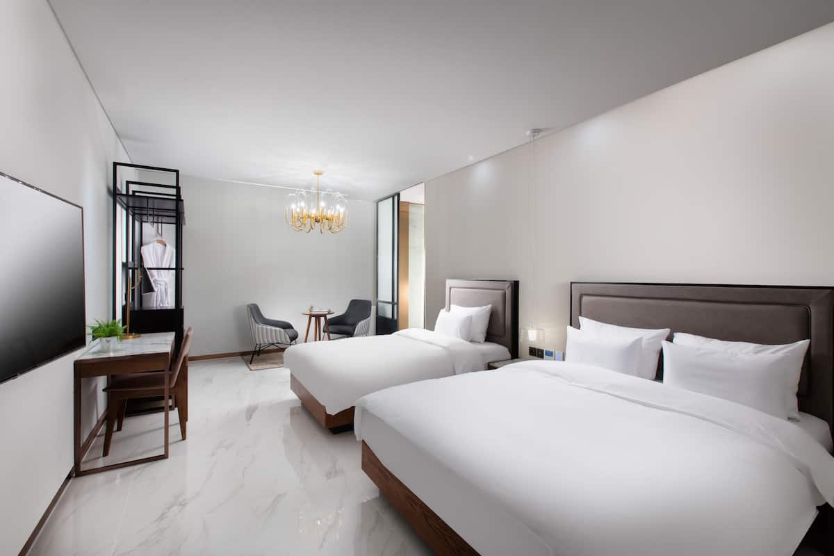 A modern hotel room with two beds, a desk, a chair, a small table, and a chandelier.