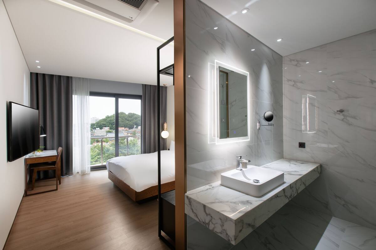 A modern hotel room with a large bed, a desk, a TV, and a bathroom with a marble sink and shower.
