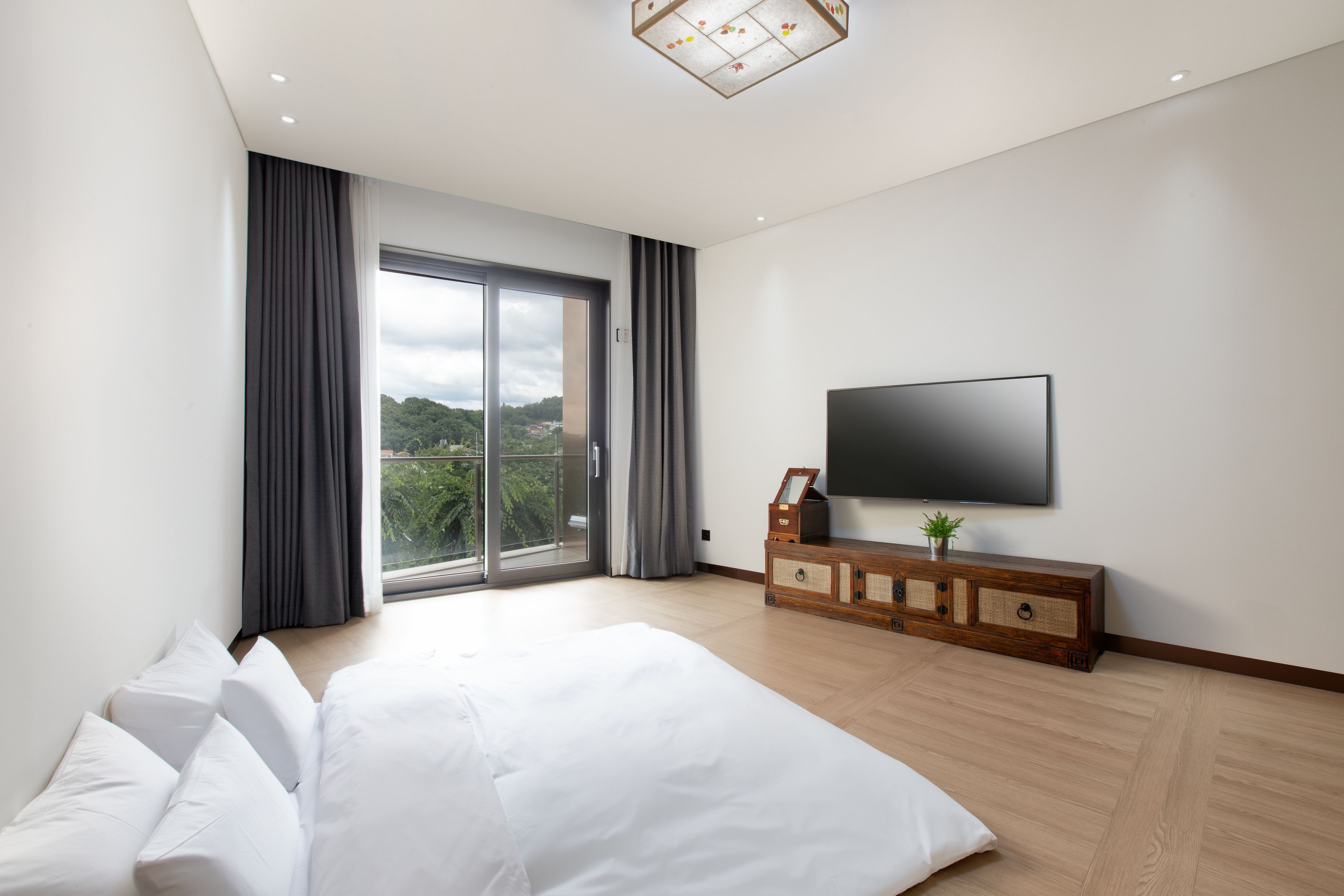 Ein modernes Schlafzimmer mit einem großen Bett, einem Fernseher auf einer Holzkommode und einem Balkon mit Vorhängen.