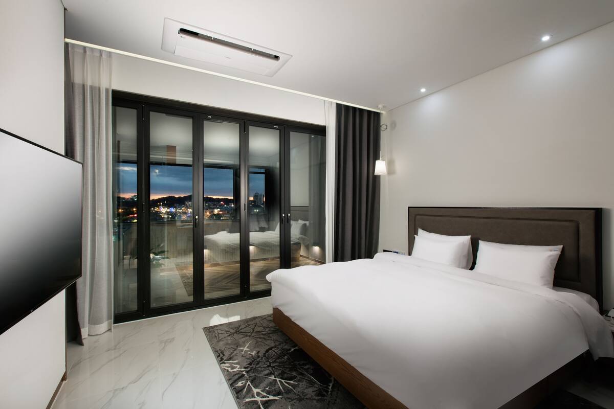 A modern hotel room with a large bed, a flat-screen TV, a balcony with a city view, and a sliding glass door.