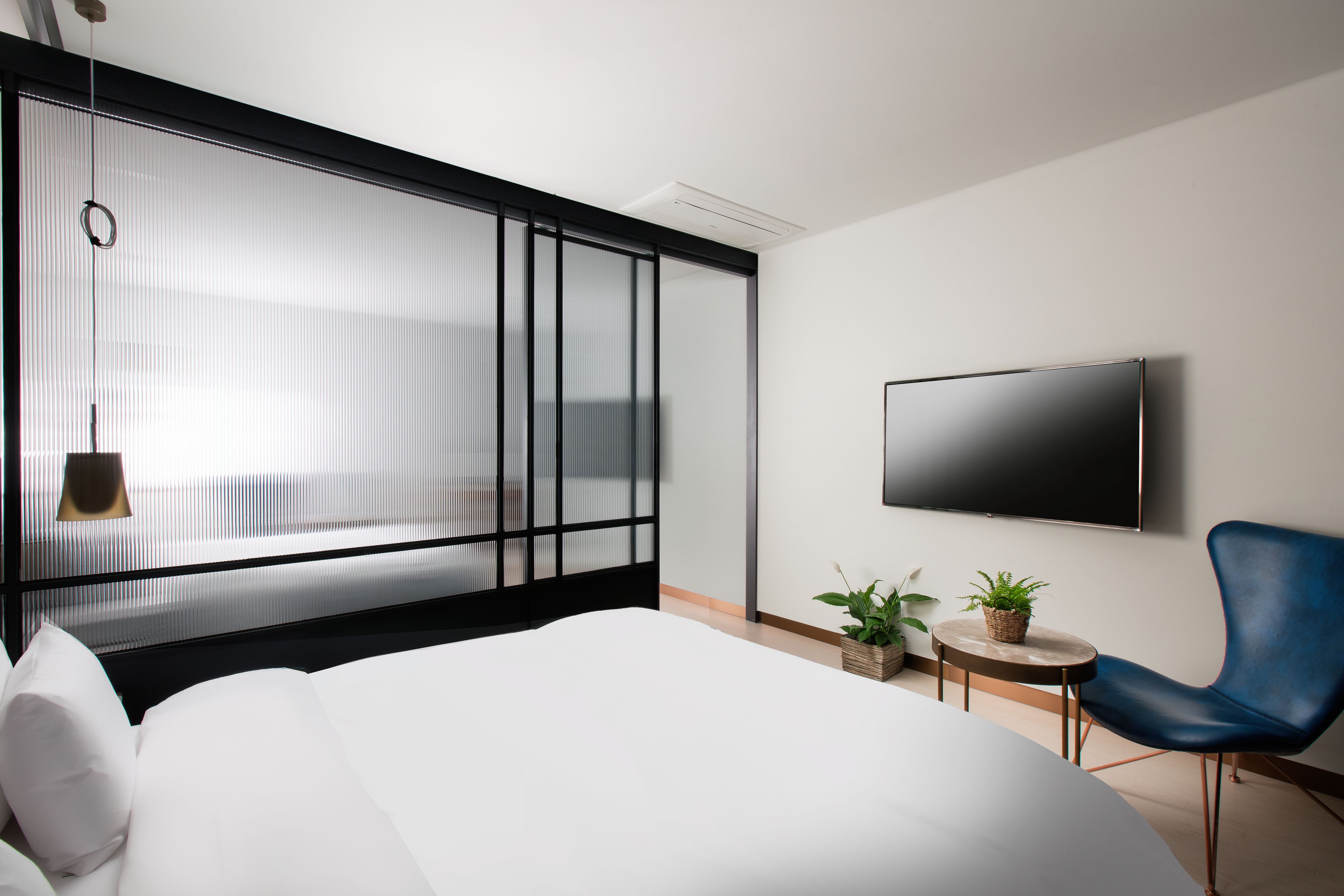 A modern hotel room with a large bed, a TV mounted on the wall, a blue chair, and two potted plants.