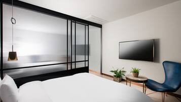 A modern hotel room with a large bed, a TV mounted on the wall, a blue chair, and two potted plants.