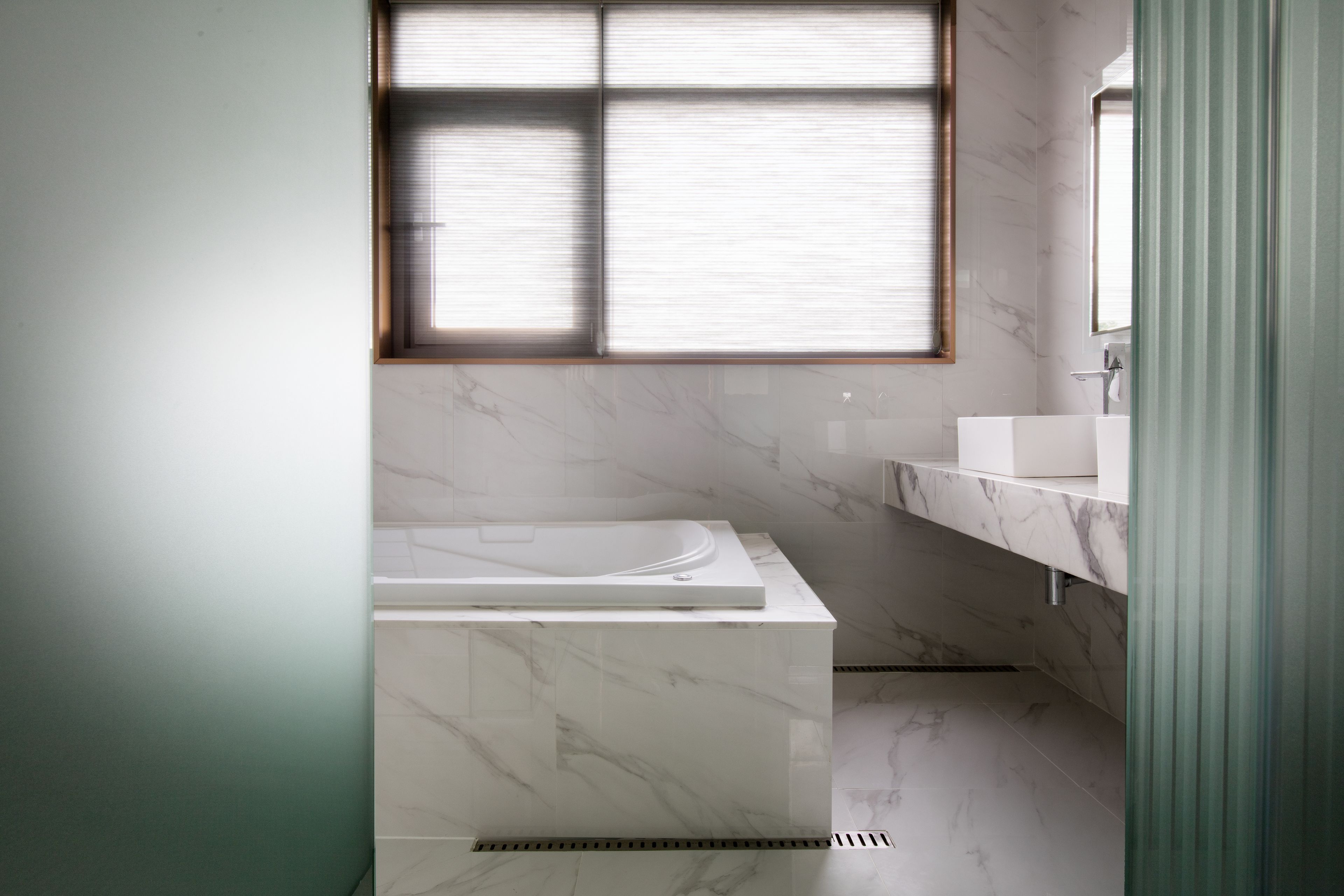 A modern bathroom with a marble sink, a bathtub, and a window with blinds.