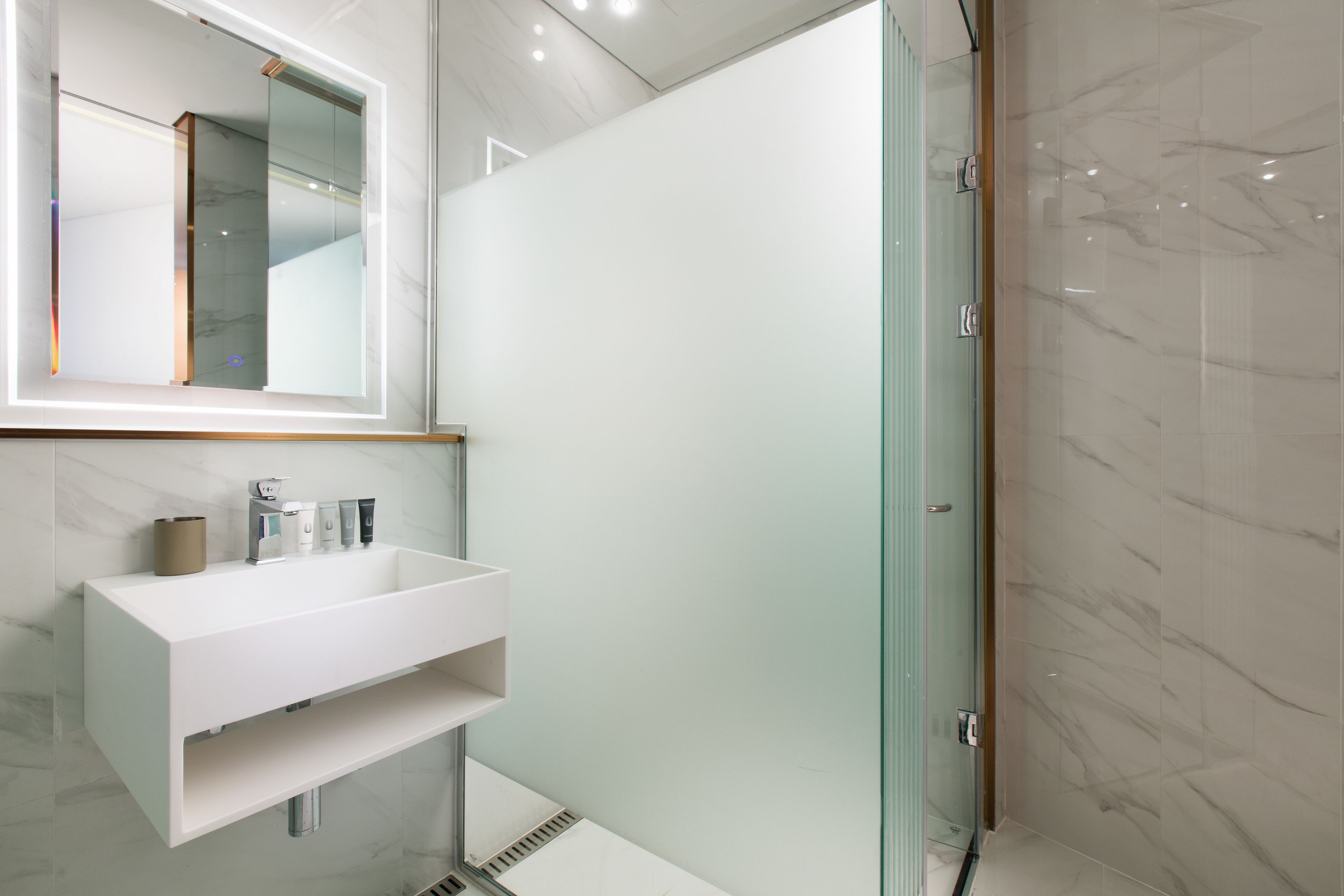 Un baño moderno con un tocador blanco, cabina de ducha de vidrio y pared revestida de mármol.