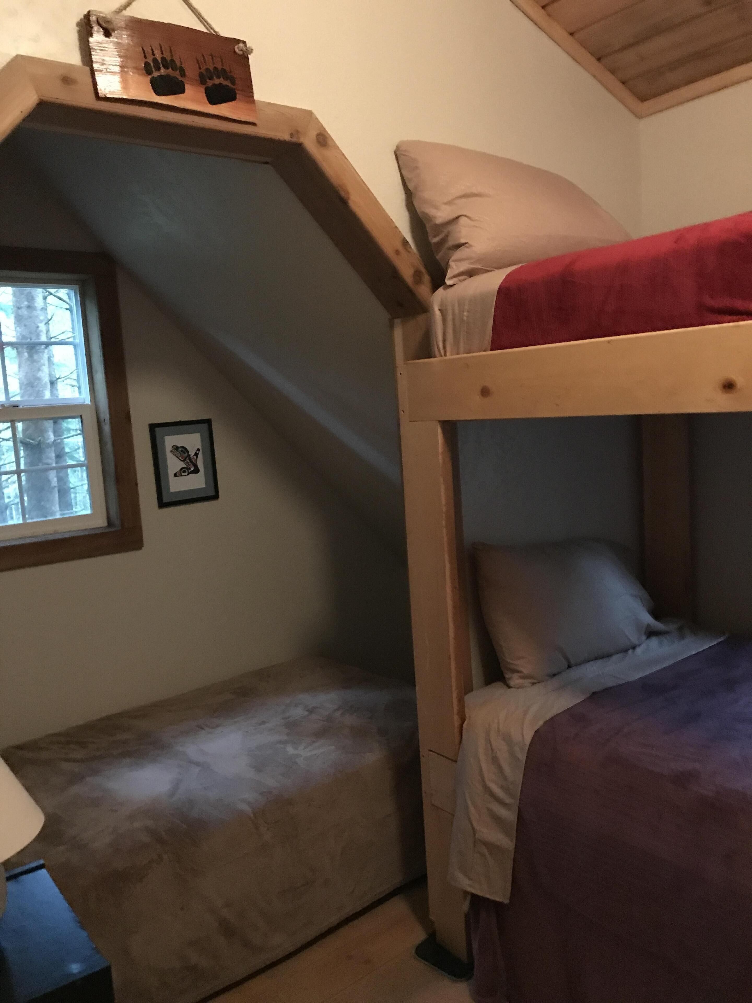 A bunk bed with a red blanket, a window with a view of trees, and a framed picture on the wall.