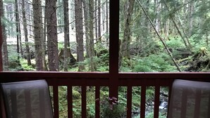 A covered porch with two chairs and a table, overlooking a forested area with a small waterfall. - Experience Alaska with a touch of home! (Hollis)