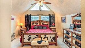 Une chambre avec un plafond en pente, un lit doté d’une tête de lit en peau de vache, une commode en bois et un ventilateur de plafond.
