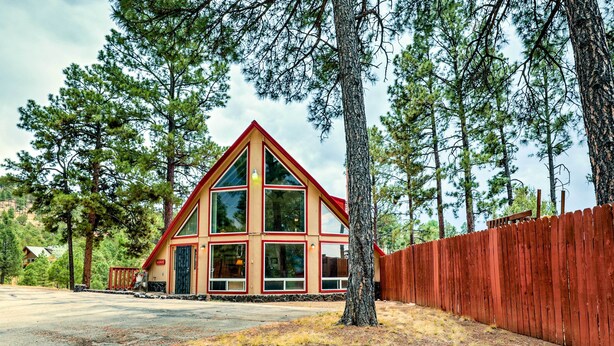 Une cabane avec un toit rouge et de grandes fenêtres, entourée d’arbres et d’une clôture en bois.
