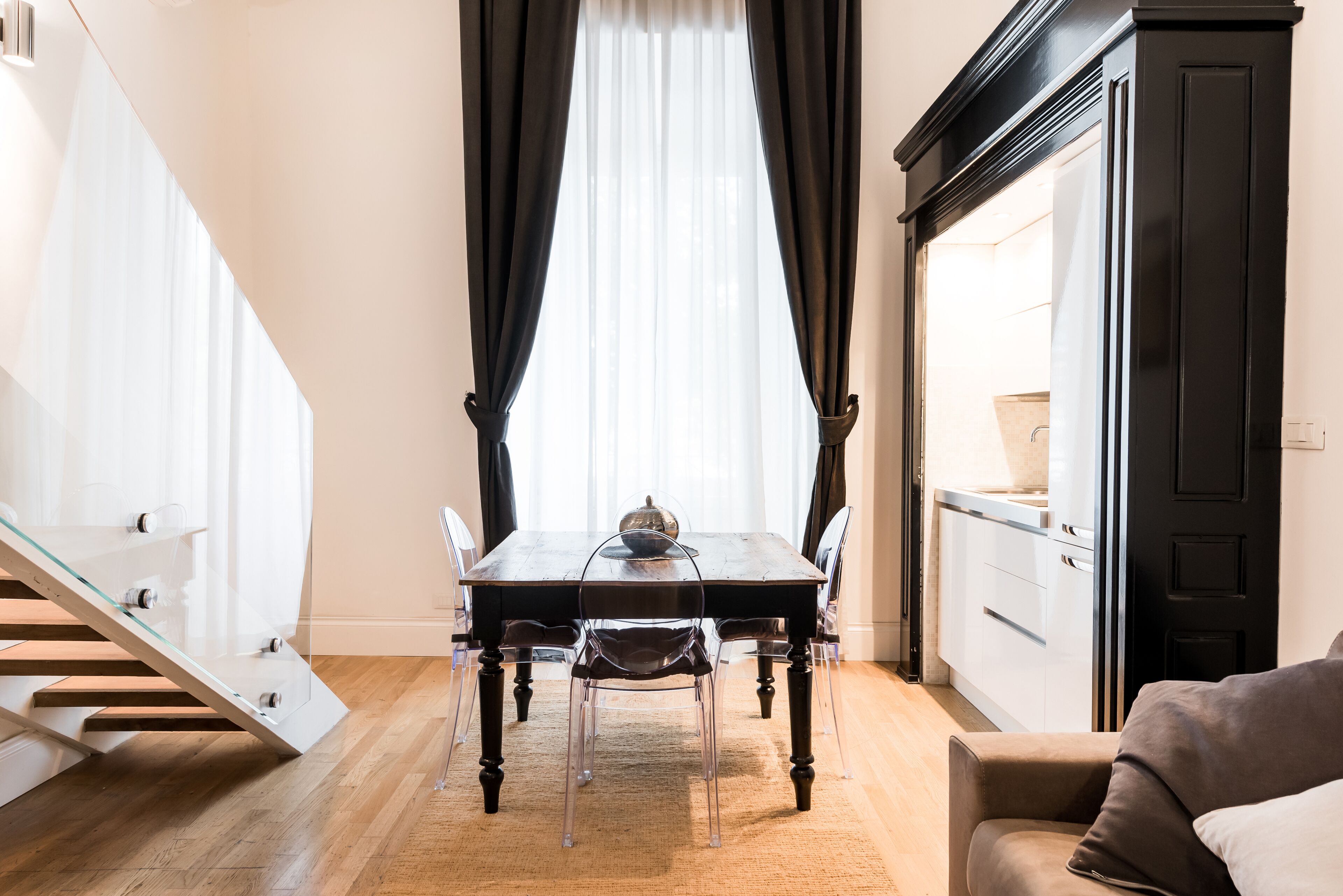 Un comedor con mesa de vidrio, sillas, una escalera y una zona de cocina.