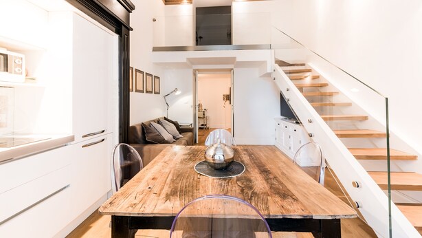 Une cuisine moderne avec des meubles blancs, une table à manger en bois et un escalier en verre menant à l’étage supérieur.