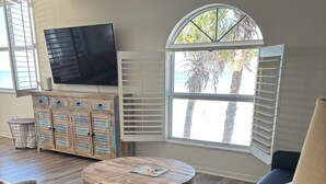 Un salon avec un téléviseur, une table basse ronde en bois, un canapé et une fenêtre avec des stores.
