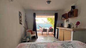 A compact bedroom with a bed, a small desk, a chair, and a view of an outdoor area with a table and chairs. - 'Boutique' studio 'Dushi Bida' (Willemstad)