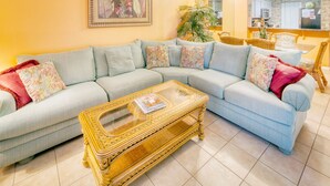 A living room with a light blue sofa, floral cushions, a wicker coffee table, and a framed floral painting on the wall. - CRC 2106 - Ocean and Pool View Ground Floor Condo (St Augustine)
