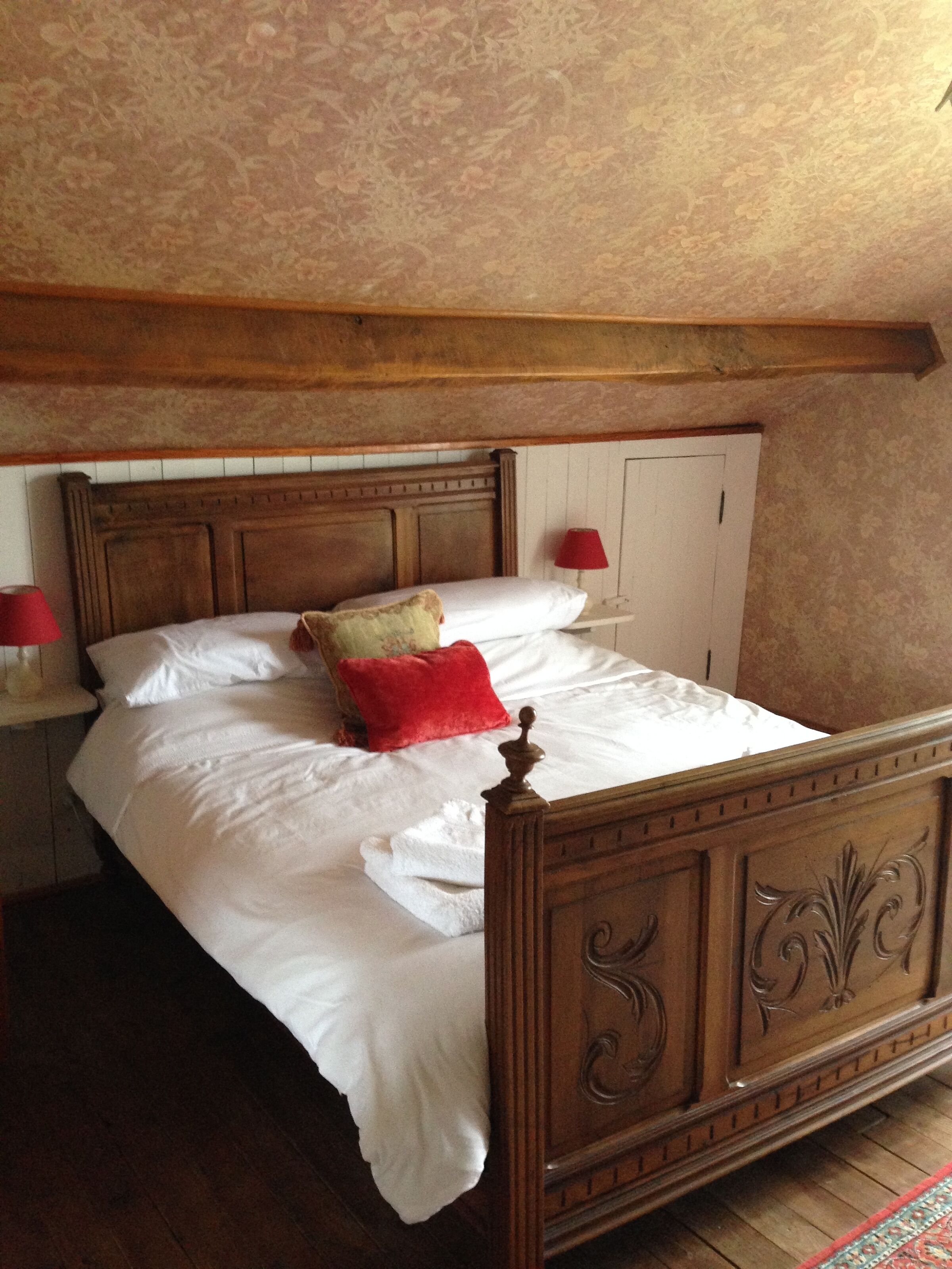 Un lit à baldaquin en bois, avec du linge de lit blanc et un oreiller rouge, une table de chevet avec une lampe rouge et un plancher en bois.