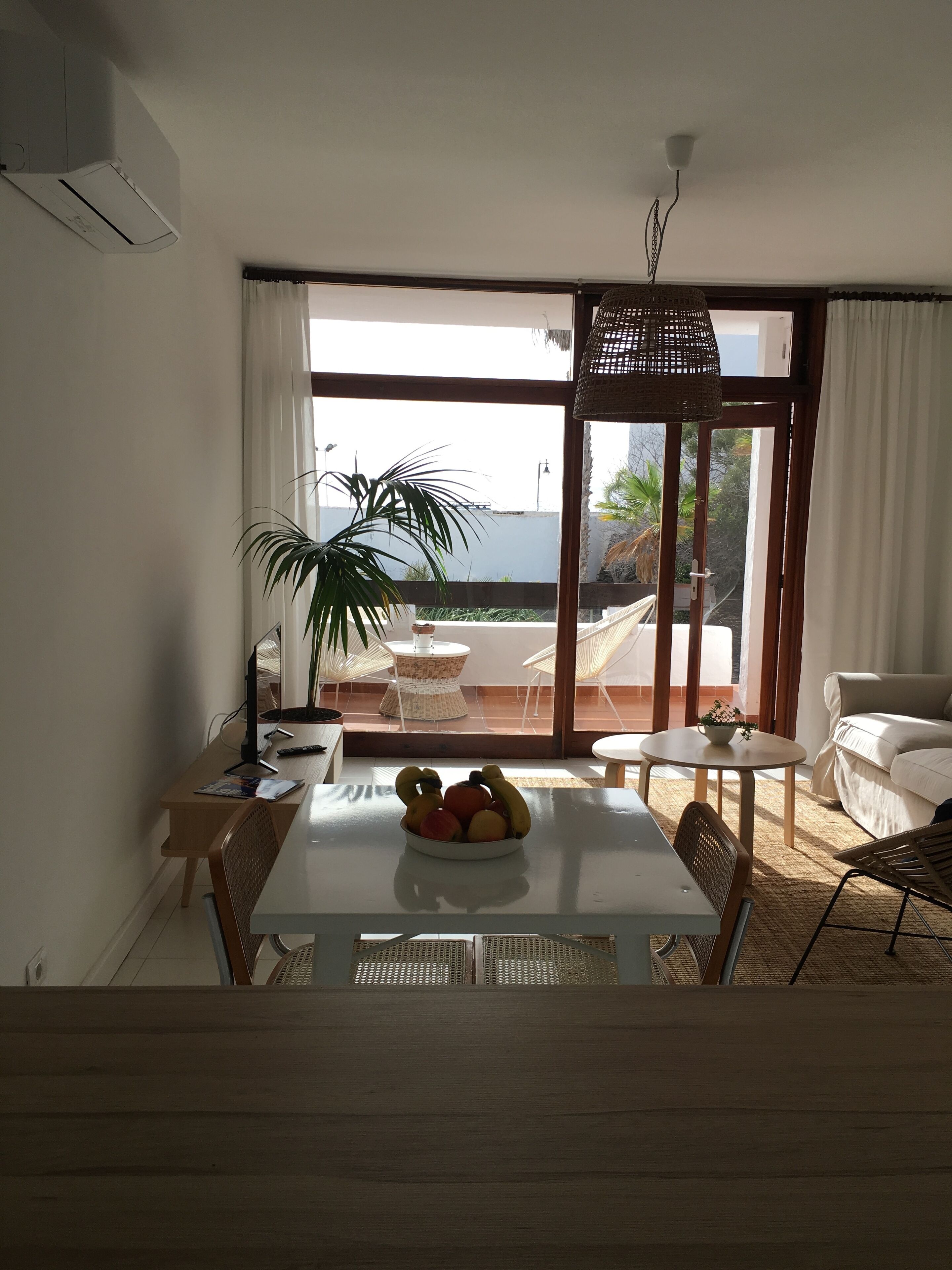 A dining area with a table, chairs, and a bowl of fruit. There is a large window with curtains, a potted plant, and a view of the outdoors.