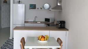 Una cocina moderna con armarios blancos, una campana extractora de acero inoxidable, un microondas y una cafetera sobre la encimera. Hay una mesa de comedor con un cuenco de fruta y dos sillas de mimbre.
