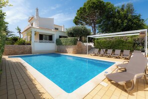 Una piscina con sillones de descanso, un muro de piedra y un edificio blanco con tejado de baldosas.