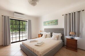 A bedroom with a bed, a nightstand, a lamp, a painting, and a view through a large window. - Villa Ladeira - Private pool, Sea view, Smart tv, games, wifi, Ac (Albufeira)