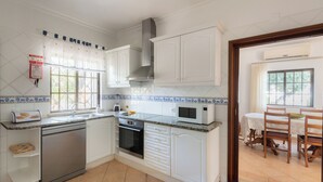 A kitchen with white cabinets, a dishwasher, an oven, a microwave, and a dining area with a table and chairs. - Villa Ladeira - Private pool, Sea view, Smart tv, games, wifi, Ac (Albufeira)