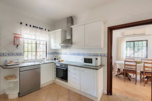 A kitchen with white cabinets, a dishwasher, an oven, a microwave, and a dining area with a table and chairs. - Villa Ladeira - Private pool, Sea view, Smart tv, games, wifi, Ac (Albufeira)