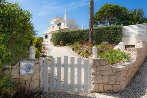 Une villa blanche, dotée d’un mur et d’une porte en pierre, entourée de verdure et bordée d’un chemin pavé.