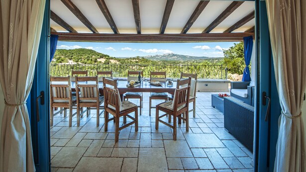 An open patio with a wooden table and chairs, a wicker sofa, and a view of a hilly landscape.