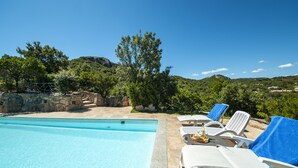 A pool area with clear blue water, white lounge chairs with blue towels, and a view of green hills and trees.