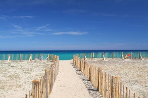Un sentier de sable qui longe la plage et mène à l’océan, bordé de clôtures en bois et agrémenté d’un parasol de plage.