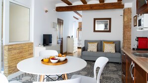 A modern kitchen with a dining area, a grey sofa with patterned cushions, and a television in the background.