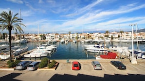Un puerto deportivo con barcos amarrados, un aparcamiento con coches y un pueblo costero al fondo.