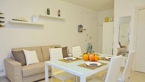 A modern living room with a white sofa, a dining table set for two, and a kitchen area with a refrigerator and shelves. - House in front of the beach of Balestrate (Balestrate)
