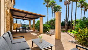 A covered patio area with outdoor furniture, a table with a vase, and a view of palm trees and greenery. - Son Sastre 024 luxurious villa with large swimming pool, air conditioning, garden and terrace (Muro)