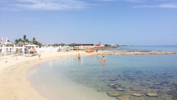 A beach with clear shallow waters, umbrellas, and a few people.