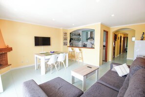 Ein Wohnzimmer mit einer Couch, einem Couchtisch und einem Fernseher. Es gibt eine Küchenzeile mit einer Arbeitsplatte und Stühlen.