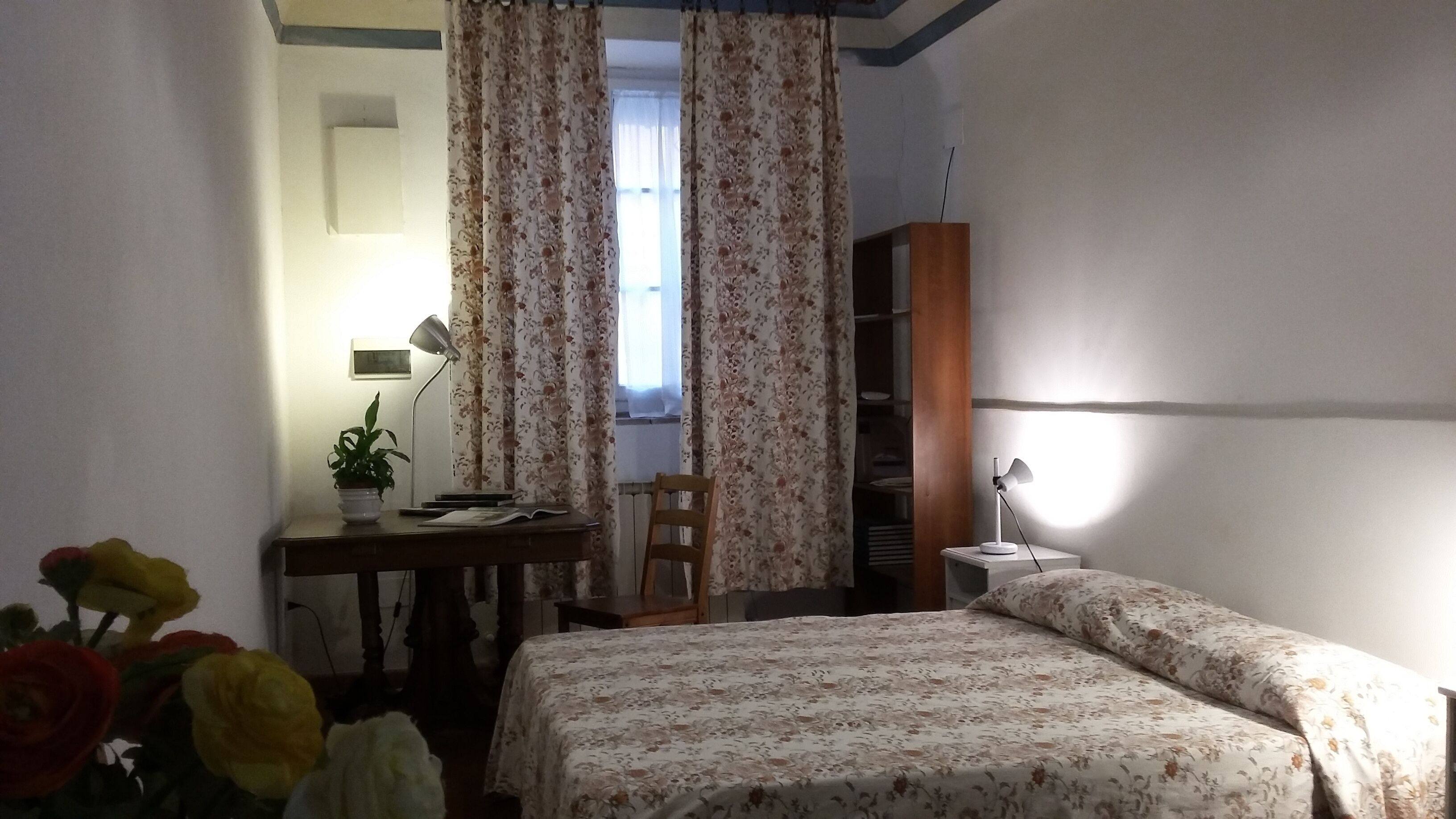 A bedroom with a bed, a desk with a lamp, a chair, a radiator, and a wardrobe.