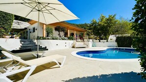 Ein Poolbereich mit Sonnenschirm, Liegestühlen und einer Treppe, die zu einer überdachten Terrasse führt.