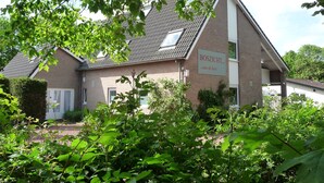 A two-story building with a sloped roof, a sign reading "BOSZICHT... aan de kust," and a garden area in front.