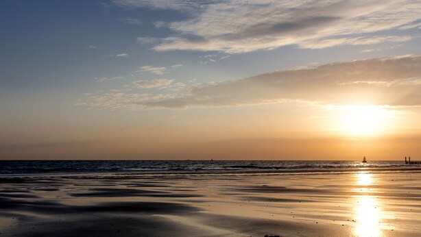 A beach with a sunset, calm sea, and a distant lighthouse.