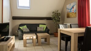 A modern living room with a black leather sofa, two wooden side tables, a dining table with chairs, a potted plant, and a painting of a lighthouse.