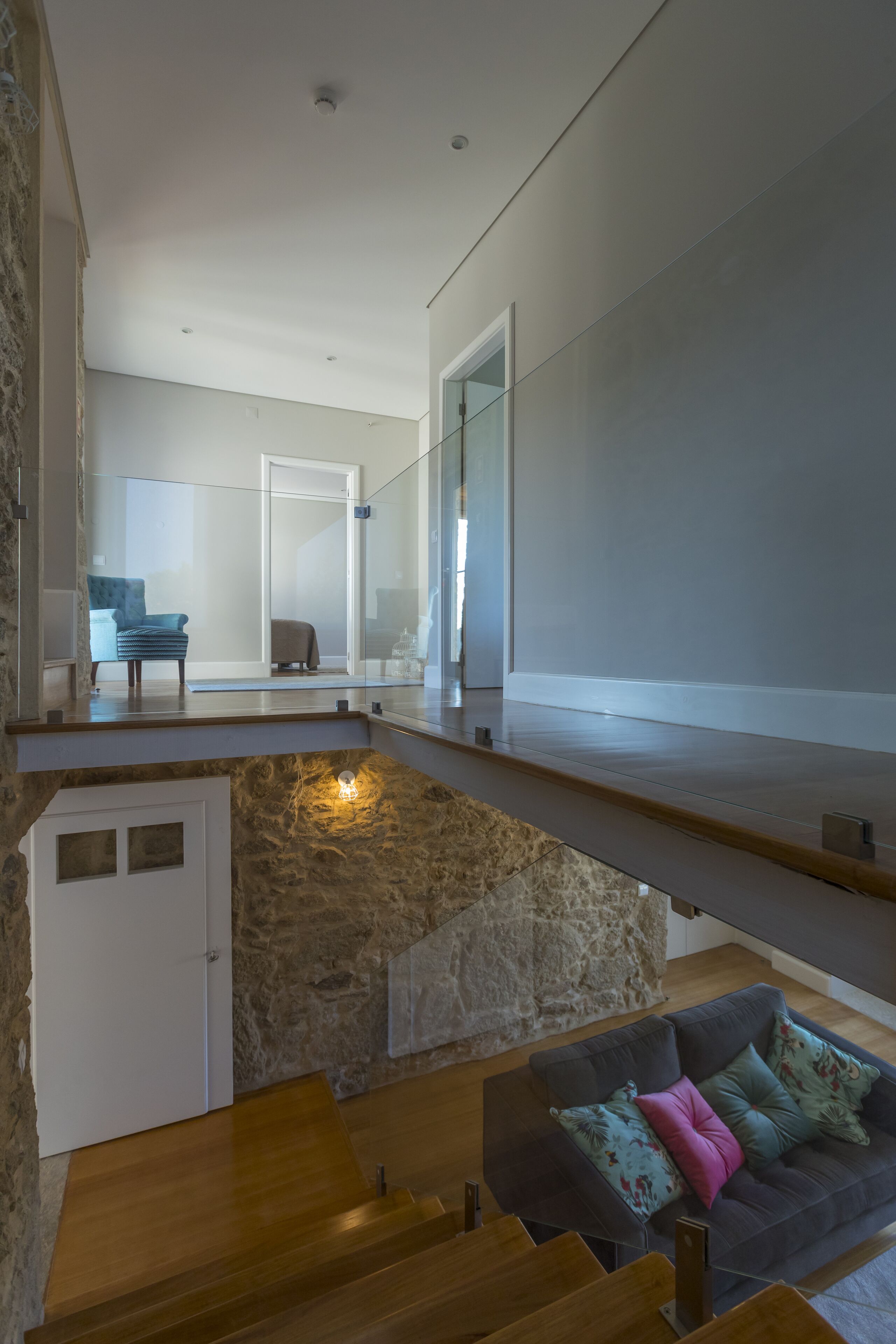 Un intérieur moderne avec un mur en pierre, un escalier en bois, un canapé gris agrémenté de coussins à motifs et une porte en verre donnant sur une autre pièce.
