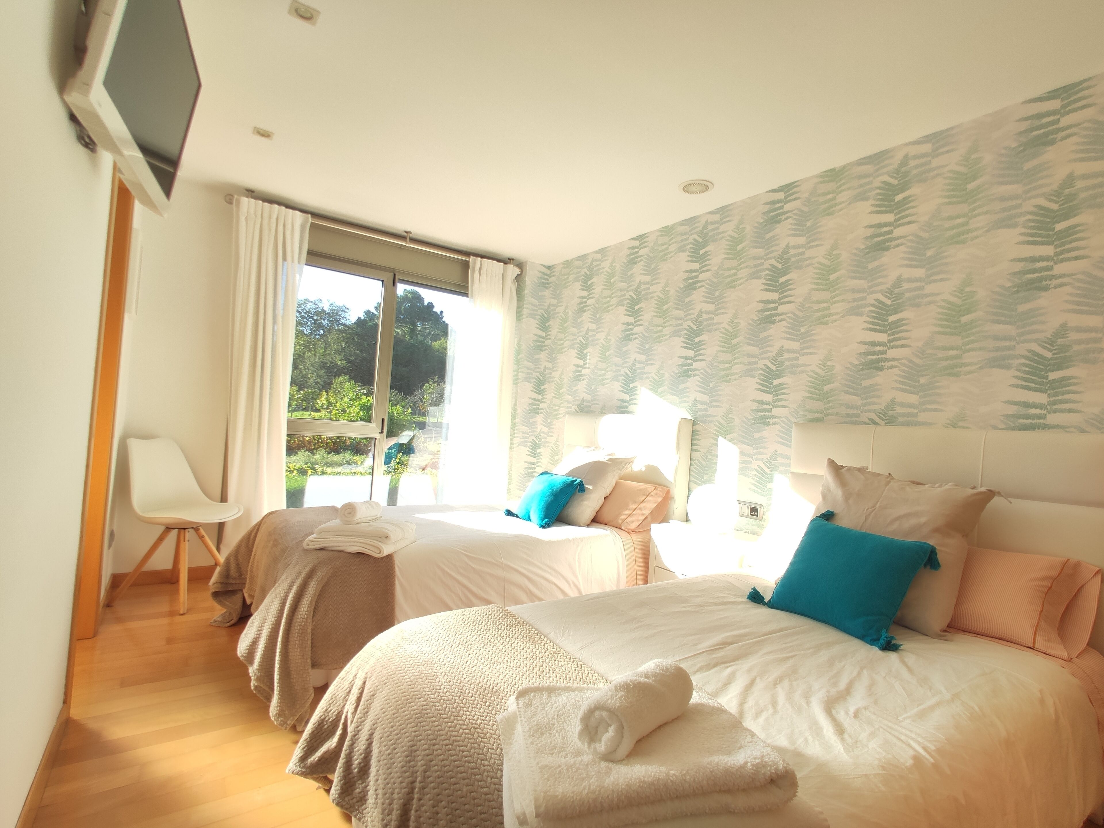 A hotel room with two beds, a TV, a chair, and a window with a view of greenery.