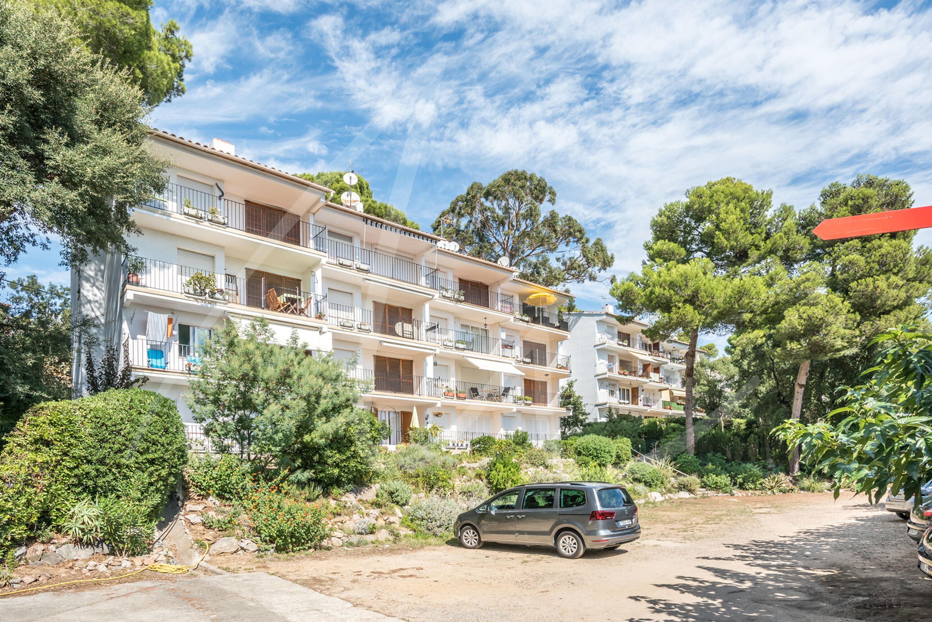 A multi-story residential building with balconies, surrounded by greenery and parked cars.