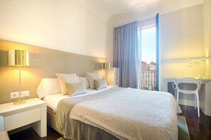 Hotellihuone, jossa on suuri sänky, työpöytä tuolilla ja ikkunasta avautuva näkymä rakennuksiin.
