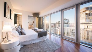 Ein modernes Schlafzimmer mit einem großen Bett, einem Nachttisch mit Lampe und Blick auf die Stadt durch raumhohe Fenster.
