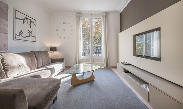 Una sala de estar moderna con un sofá, una mesa de centro de vidrio, un televisor montado en la pared y un ventanal con cortinas.