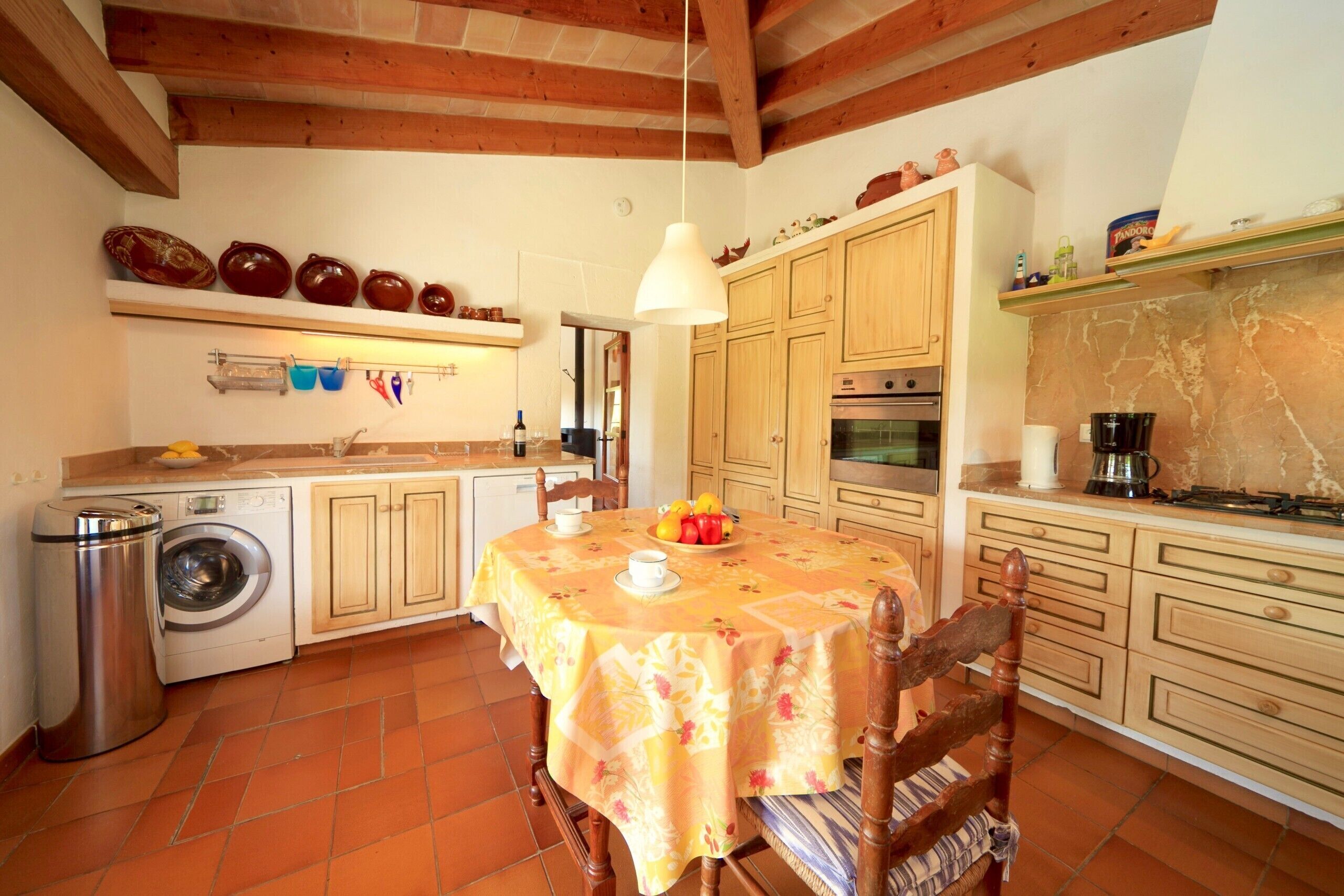 Une cuisine avec des meubles de rangement en bois, un lave-linge, une table à manger recouverte d’une nappe à motifs floraux et un luminaire suspendu.