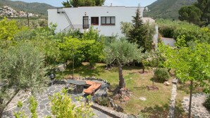 A white building with a stone patio, surrounded by greenery and hills in the background. - Sea view, terrace 20 m2, WIFI, parking, barbecue and garden of 1000 m2. (Roses, Girona)