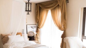 Un dormitorio con cama, una silla, una mesita de noche, una lámpara y una ventana con cortinas.